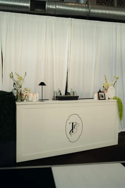 Wedding bar setup with white bar decor, monogram decal, candles, florals, glassware, and greenery garland under industrial ductwork indoors