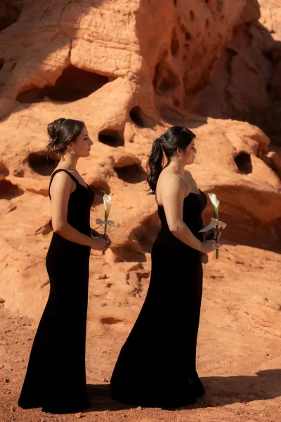 Bridesmaid portrait in black bridesmaid dresses holding white calla lily bouquets with ribbon bows, lined up against red rock desert backdrop