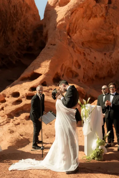 Ceremony kiss as bride and groom embrace in veil and tux by a floral podium, with red rock cliffs under a blue sky behind them