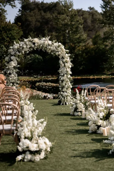 Ceremony arch, a circular floral arch, frames a pondside garden aisle flanked by bentwood chairs beneath leafy trees.
