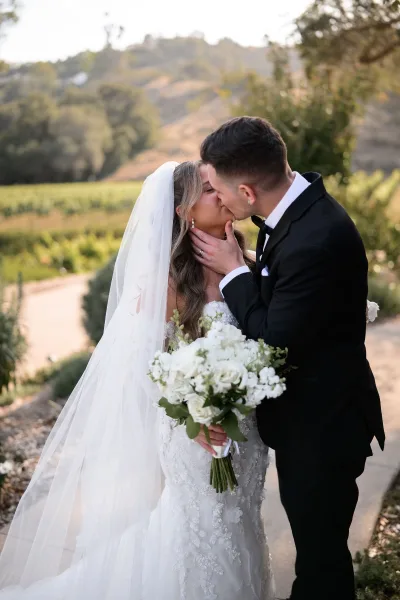 Wedding kiss portrait as the groom holds the bride's face, her long veil catching light in a vineyard among rolling hills and trees.