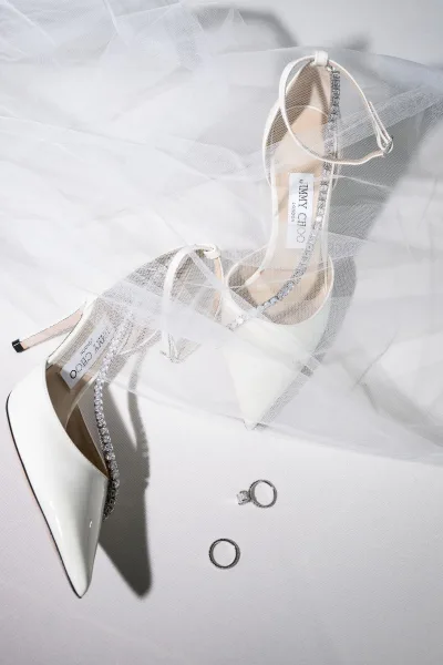 Bridal shoes, white wedding heels with crystal straps, resting on soft white fabric with gentle shadows for an elegant detail shot