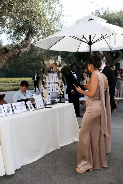 Live wedding illustrator at a wedding portrait bar sketching guests, with a floral garland accent, beneath olive trees on an outdoor patio.
