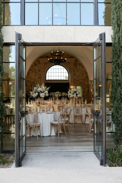 Reception decor in an indoor reception design with round tables and taper candles beneath arched windows and a stone wall backdrop.