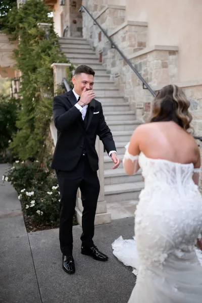 Wedding first look with the groom's surprised reaction as the bride approaches, her gown train sweeping a stone staircase with ivy and garden roses.