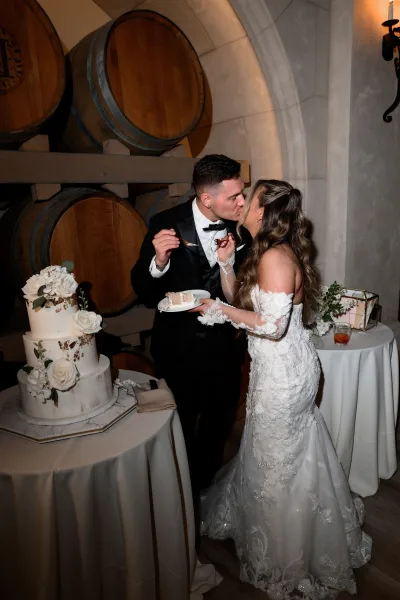 Cake cutting moment with a couple feeding cake beside wine barrels and a stone wall, the three-tier cake trimmed with white flowers.