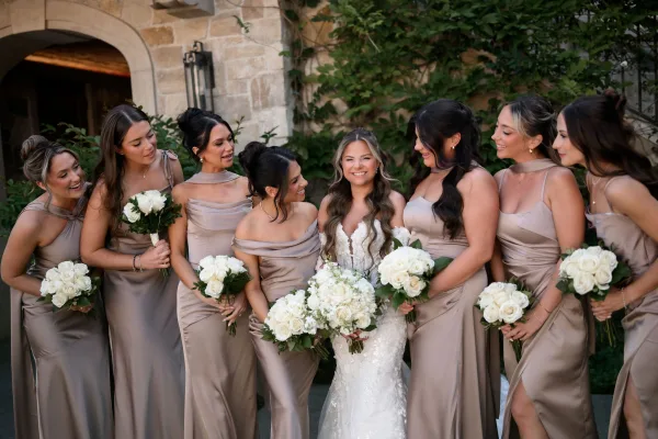 Bridal party portrait with bridesmaids holding bouquets, the bride among champagne satin dresses and white rose bouquets by an ivy-clad stone arch.