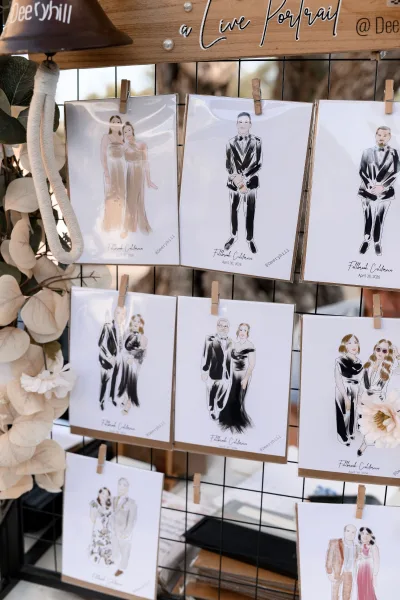 Guest portrait station for live wedding portraits, featuring illustration prints beside a wooden sign in an outdoor setting with foliage.