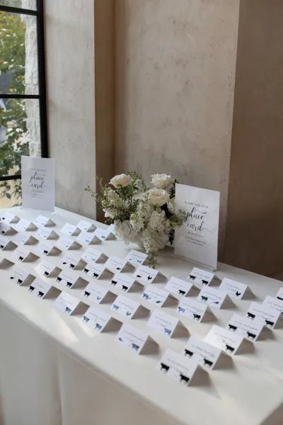 Place card display of wedding place cards arranged on table linen with white roses, set before a window framing outdoor greenery.