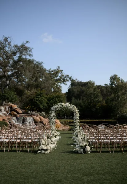 Ceremony setup with a floral wedding arch on a grass lawn, overlooking a waterfall cascading over rocks into a pond, framed by trees.