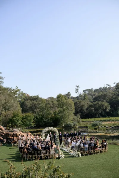 Outdoor ceremony setup for a garden wedding ceremony featuring a floral arch on a lakeside lawn with trees and hills.