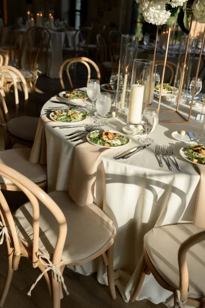 Reception tablescape on a round wedding table featuring a white hydrangea centerpiece in a candlelit room with stone walls and surrounding tables.
