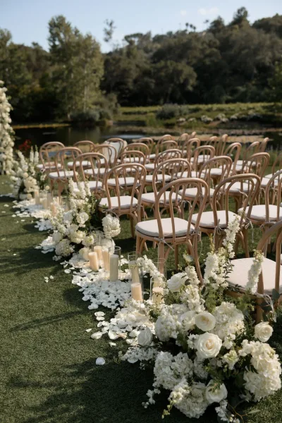Ceremony aisle design featuring white roses, bordered by outdoor ceremony seating on a lakeside lawn with trees and a distant hillside.