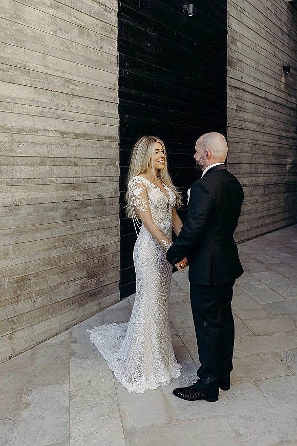 Couple portrait with bride and groom holding hands, bride gazing at bald groom in black tux beside a concrete and black wood wall backdrop