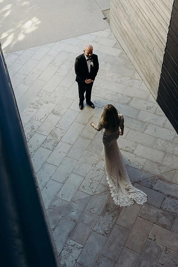 First look moment as bride approaches groom in tuxedo, lace train and veil trailing across stone tiles in a modern courtyard