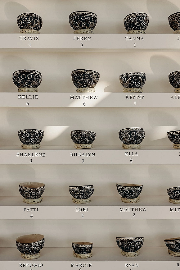 Wedding seating chart escort card display with place cards in patterned bowls tied with raffia on shelves against a white wall backdrop