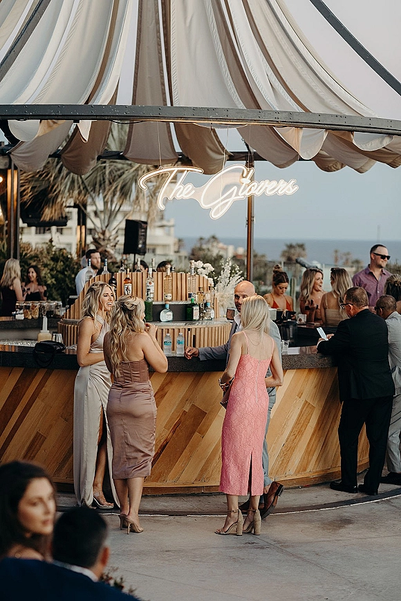 Wedding cocktail hour wedding bar setup under a striped canopy, with neon name sign, florals and bottles on a patio with ocean view and palms