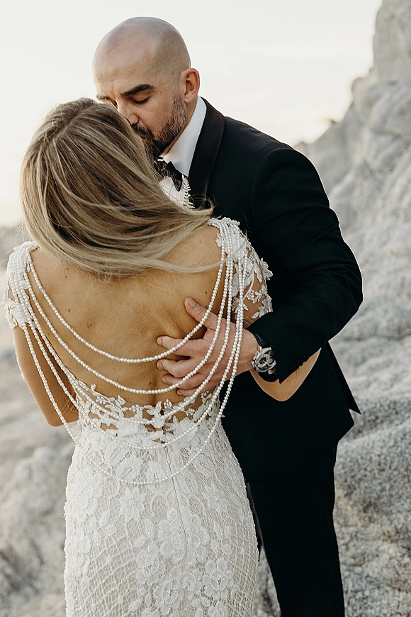 Wedding couple portrait of groom kissing bride’s forehead as they embrace, her open-back lace dress with pearl straps on ocean cliffs
