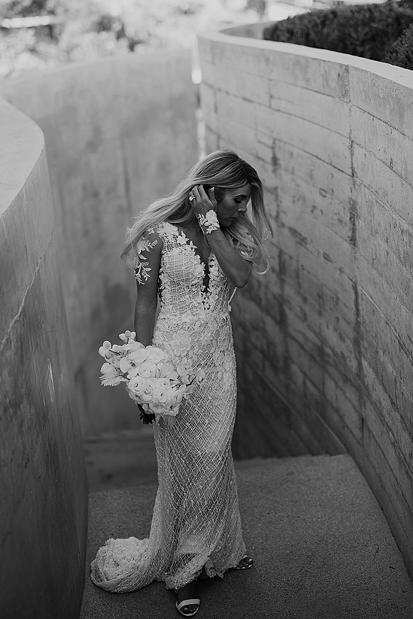 Bridal portrait of a bride in a long sleeve lace wedding dress holding a white bouquet on concrete stairs, looking down at her ring