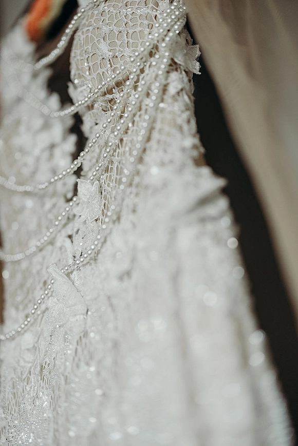 Wedding dress close-up showing lace wedding dress detail with pearl strands draped over floral lace appliques on soft neutral fabric background