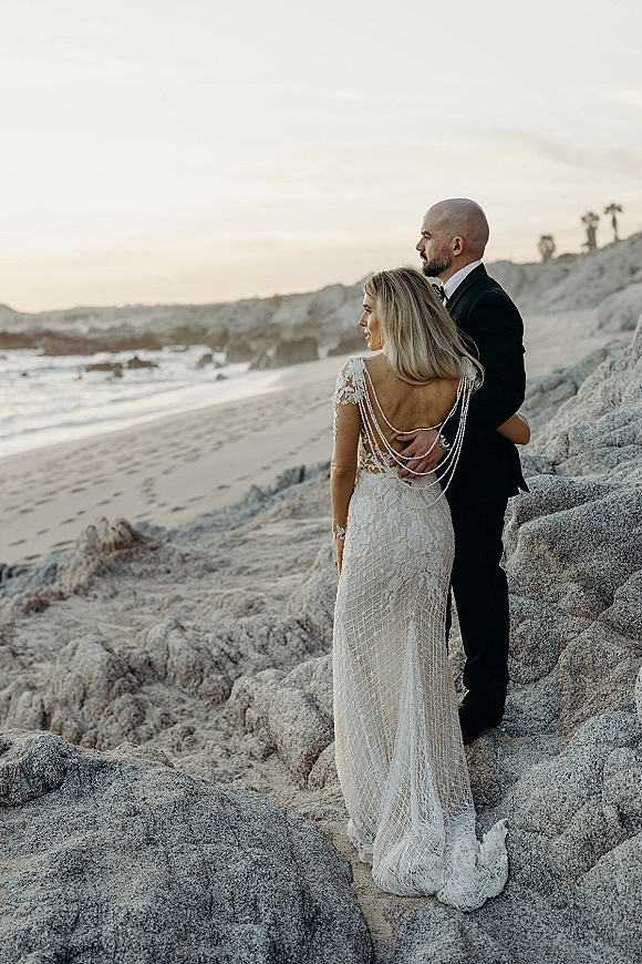 Couple portrait of bride and groom from behind on a rocky beach at sunset, lace open-back gown with pearl drape and train by waves