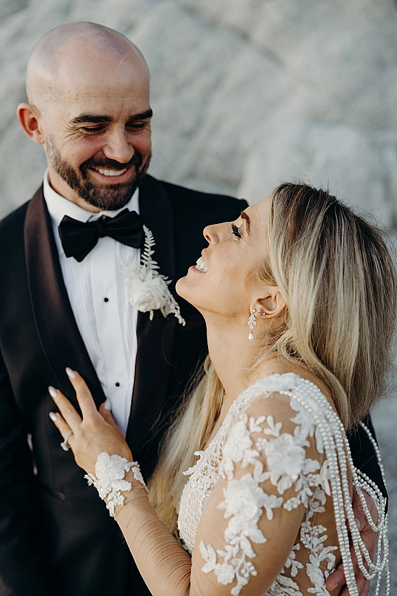 Couple portrait of bride and groom smiling, bride holding his lapel in black tuxedo and lace-sleeve gown against stone wall