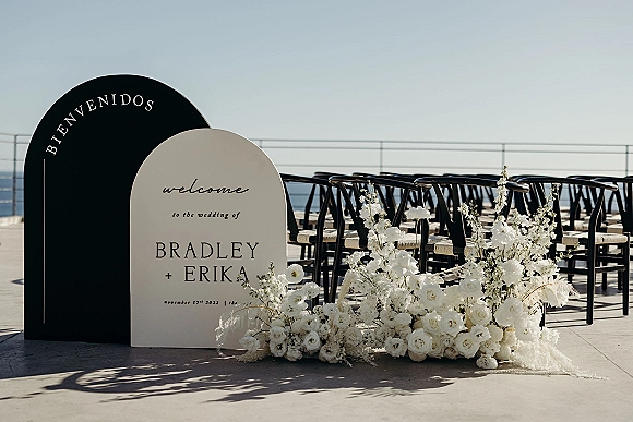 Wedding welcome sign with an arched wedding sign and white rose florals on a stone terrace, black chairs and ocean horizon behind