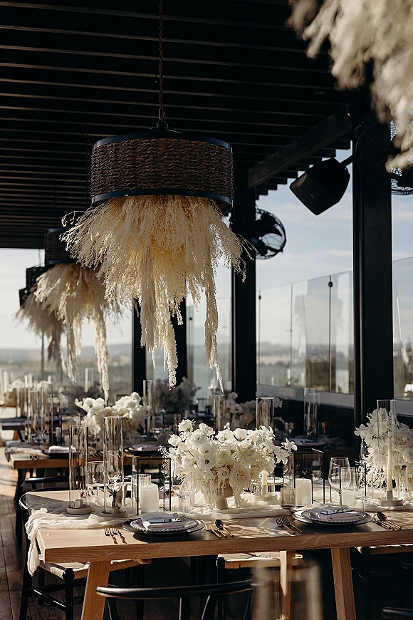 Reception tablescape with white flower centerpieces and pillar candles on wooden tables, beneath woven pendant lights by glass walls with skyline view