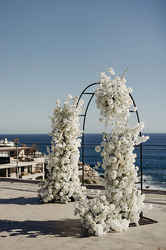 Wedding ceremony arch with white floral arch wedding arrangements on a black metal frame, set on a rooftop terrace overlooking the ocean