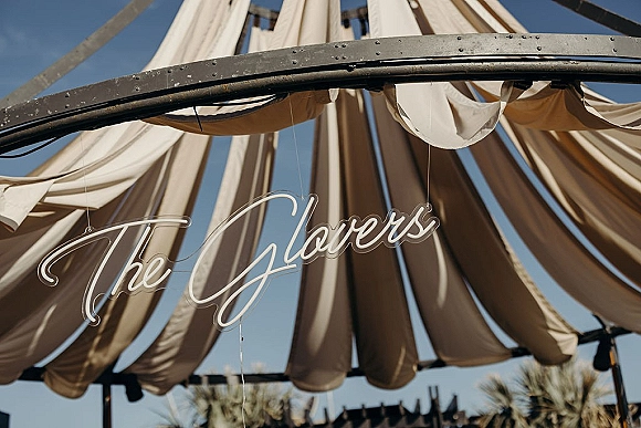 Wedding neon sign glowing in cursive, custom wedding neon sign hung from a metal frame with draped fabric under blue sky and trees