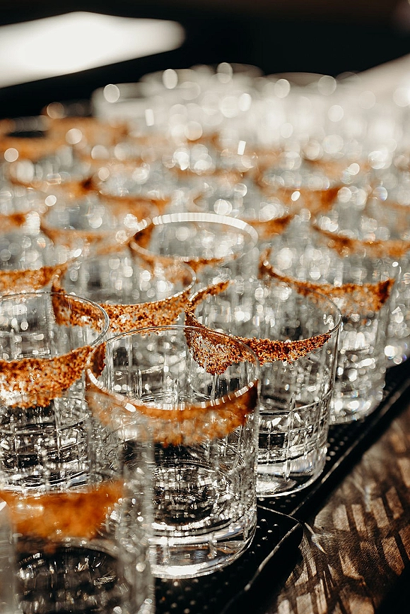 Wedding drink station with sugar rim cocktail glasses lined up beside a cocktail rimmer tray on a bar counter in dim indoor lighting