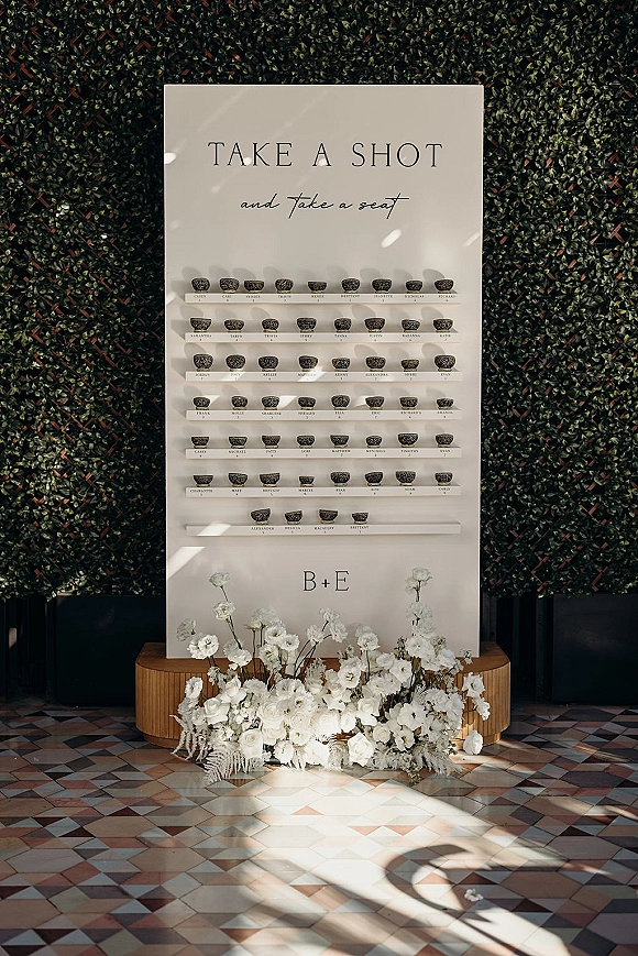 Wedding seating chart with shot glass escort cards on shelves, large printed sign with guest names, set against a green hedge wall