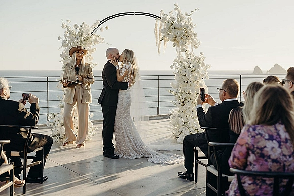 Ceremony kiss as bride and groom embrace under a white floral arch, veil flowing, with ocean cliffs behind and guests seated nearby