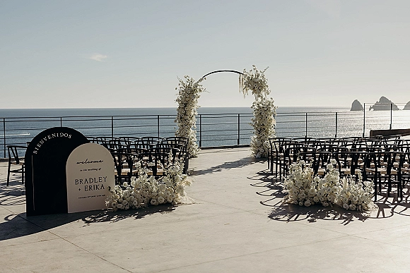 Ceremony setup with oceanfront wedding ceremony arch of white florals, black chairs lining an aisle on a terrace with ocean views