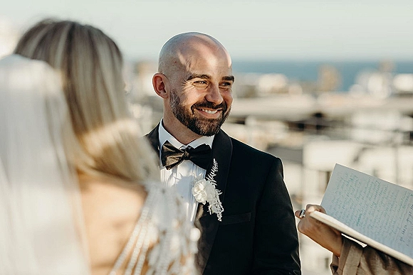 Groom portrait in a black tuxedo with bow tie and white rose boutonniere, smiling on a rooftop terrace with sky and distant buildings