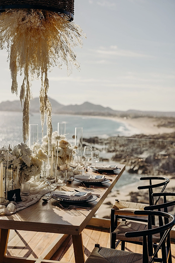 Reception tablescape with white florals and taper candles on a wood dining table, set oceanfront with rocky shoreline and mountains behind