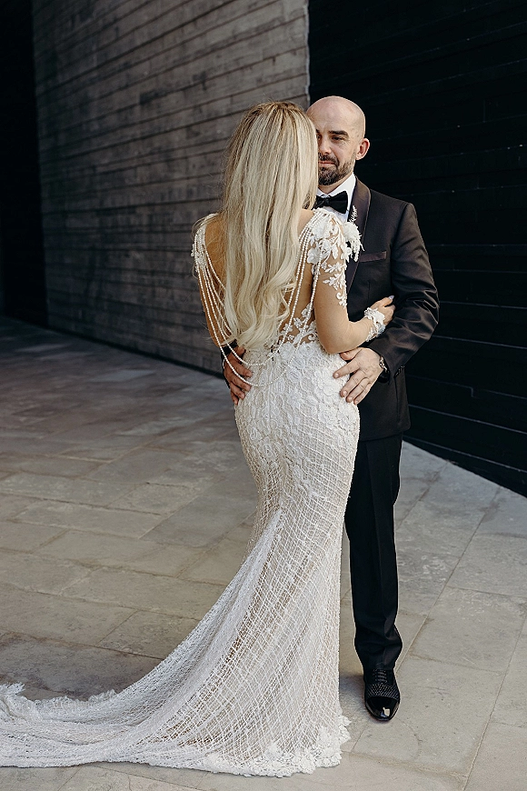 Couple portrait of bride and groom embrace, her open-back lace gown with pearl drape and long train against a dark brick wall backdrop