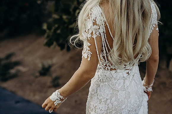 Bridal dress details with an open back lace wedding dress, illusion sleeves, and pearl back drape as the bride stands on a green path