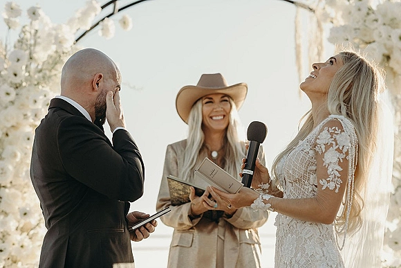 Wedding vows as the bride reading vows into a handheld microphone, groom emotional beside her under a floral arch by the water in sunlight