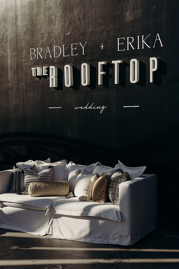 Wedding welcome sign with custom wedding wall sign 3D lettering on a dark textured wall beside a white lounge sofa in sunlight shadows