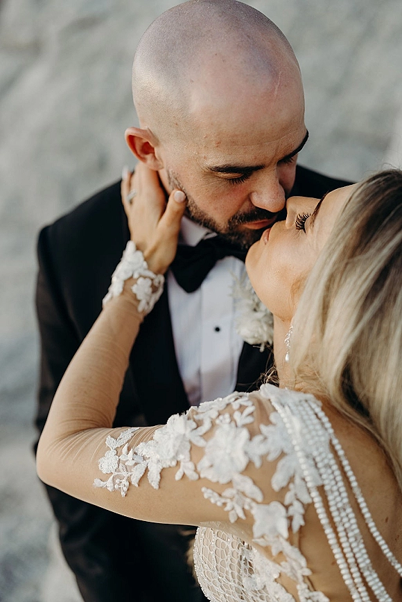 Wedding kiss portrait of bride and groom kiss, her hand on his face, lace sleeves and bow tie sharp against a gray wall