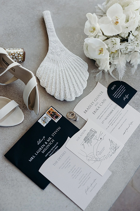 Invitation suite with black and white wedding invitations, RSVP and details cards, rings, heels, and beaded clutch on stone surface