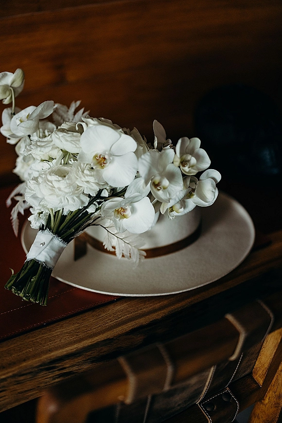 Bridal bouquet of white orchid blooms and white roses with ribbon wrap on a wooden table beside a leather suitcase and wide-brim hat
