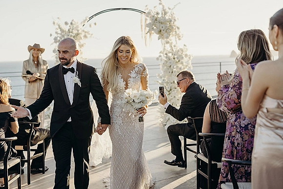 Wedding recessional as bride and groom walk hand in hand down the aisle with bouquet, guests cheering on an oceanfront terrace