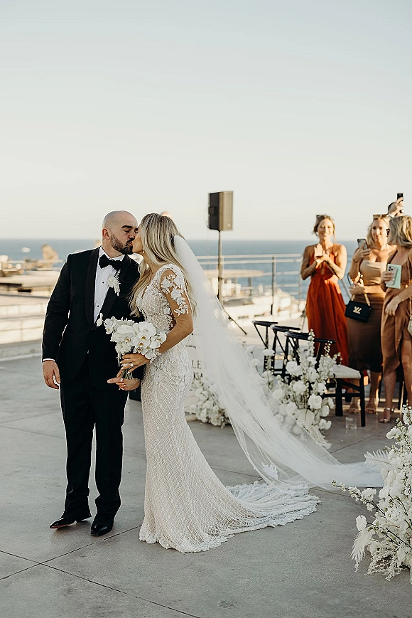 Wedding kiss as bride in lace dress and long veil holds a white bouquet, kissing groom in tux on an ocean-view terrace patio