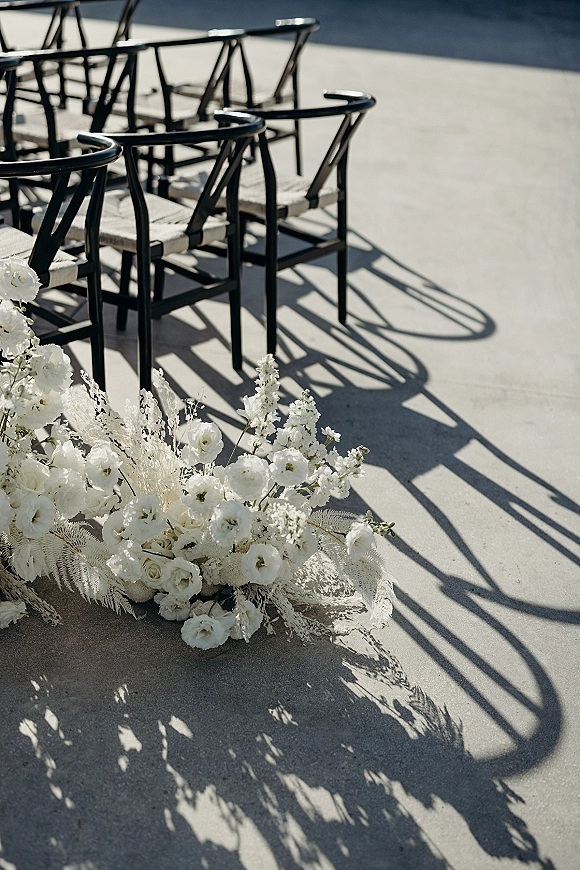 Ceremony aisle decor with white floral arrangement lining black chairs and light cushions on a sunlit concrete patio with shadows