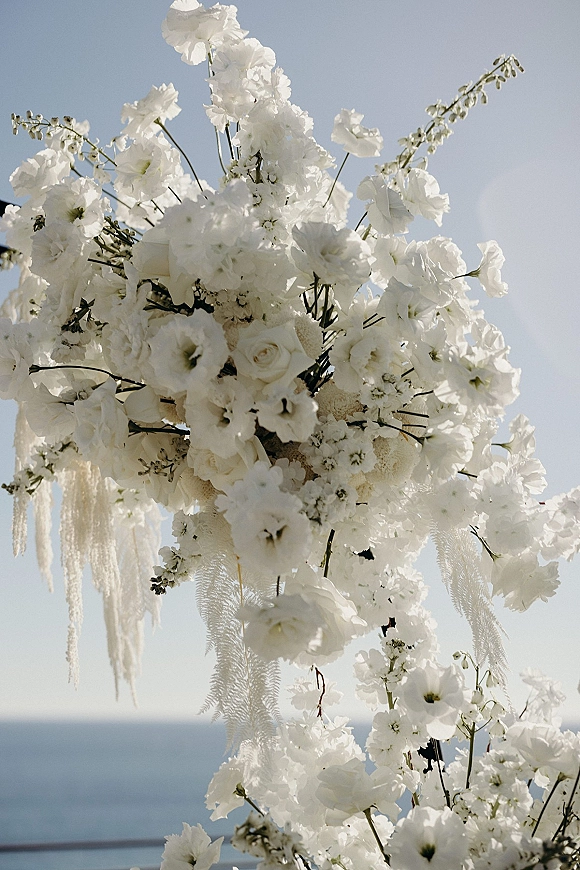 Wedding arch flowers with white roses and hanging floral strands accented by greenery branches, set against a blue sky and ocean horizon