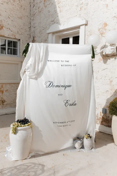 Wedding welcome sign with draped fabric, sage green ribbon ties, ceramic vases and grapes arranged on a concrete patio by a stucco wall window