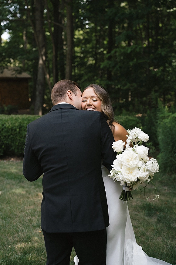 Couple portrait of bride and groom hug as he whispers to her, her white rose bouquet in hand, framed by trees and greenery outdoors