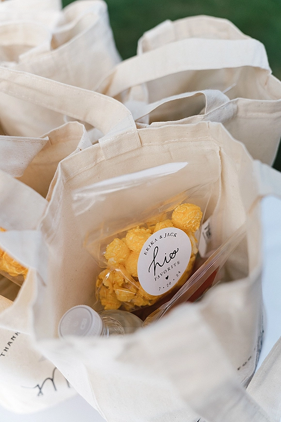 Wedding welcome bags arranged with canvas tote bags, cellophane snack packs, popcorn, bottled water, and custom favor sticker labels